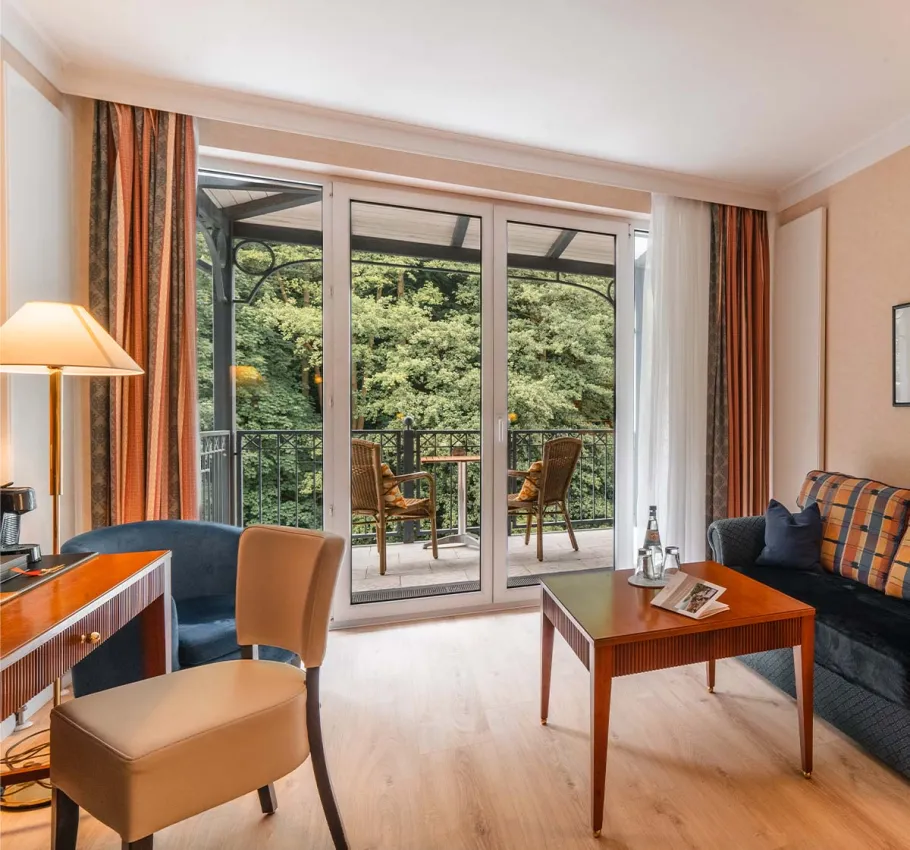 Granitz Deluxe Zimmer im Grand Hotel Binz mit großer Fensterfront, Sofa, Tisch und Balkon mit Blick ins Grüne.
