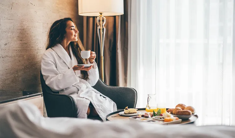 Frau im Bademantel genießt Kaffee beim Zimmerservice mit Frühstück im Hotelzimmer